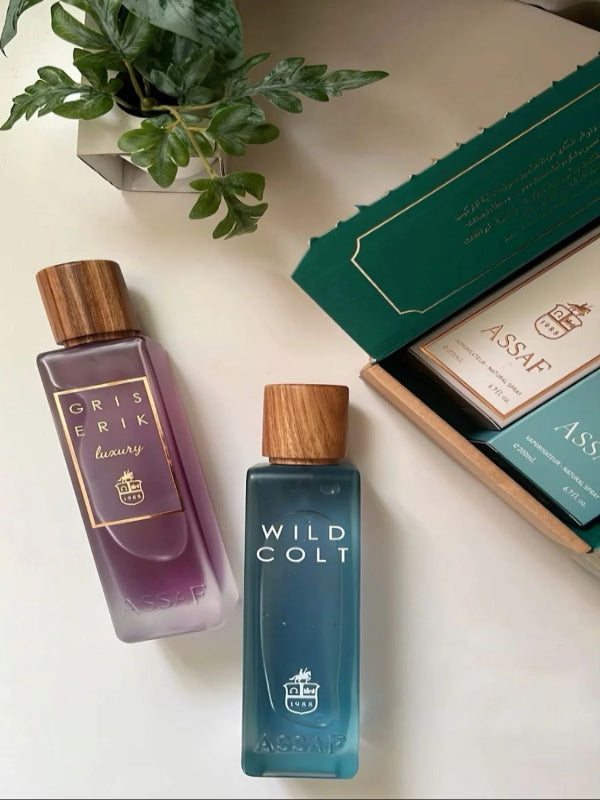 Two perfume bottles, one purple and one blue, on a white surface with a plant and box in the background.